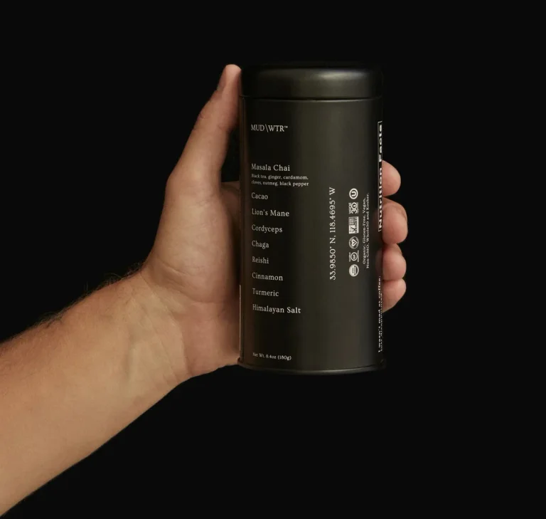 A hand holding the MUD/WTR Original cacao-and-masala-chai 180 g tin, shown in its earlier black packaging