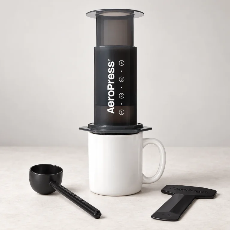 Gray AeroPress Original on a white mug beside its scoop and stirrer